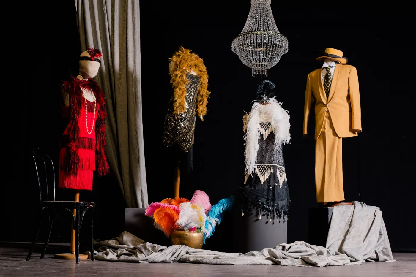Costumi anni Venti su manichini: abito rosso a frange, abiti con paillettes e boa di piume, completo color senape, sotto un lampadario di cristallo