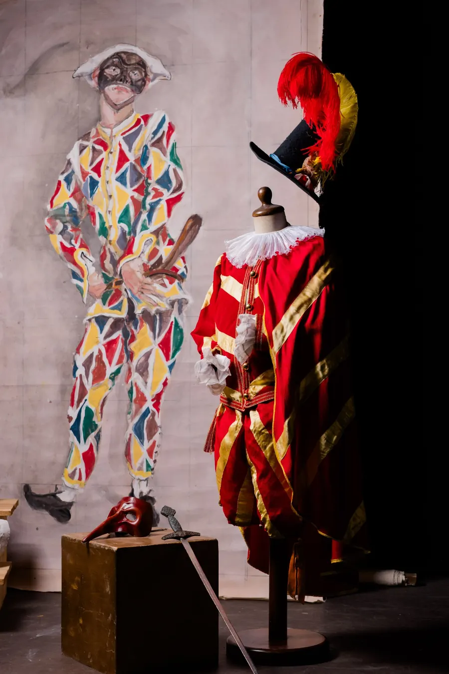 Costume di Carnevale in velluto rosso e raso oro con gorgiera e cappello piumato, davanti a un fondale dipinto con Arlecchino; accanto una maschera rossa e una spada