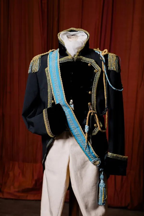 Uniforme ottocentesca blu notte con alamari dorati e fascia azzurra, davanti al sipario rosso del palcoscenico