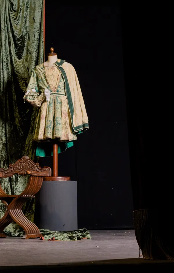 Palcoscenico allestito con una sedia dantesca intagliata, un drappo di velluto verde e un costume rinascimentale