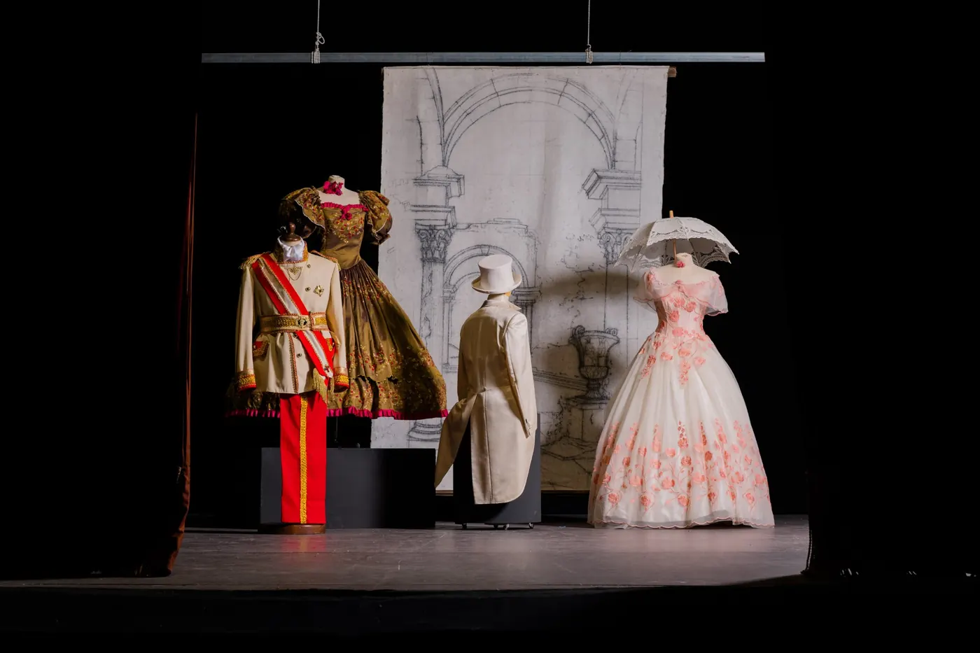 Fondale dipinto a mano con archi e colonne su un palcoscenico, davanti a costumi dell'Ottocento: uniforme con fascia rossa, abiti da ballo e frac con cilindro